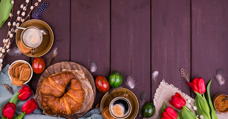 Easter breakfast with aromatic fresh espresso, croissants, brownie cake, colored eggs, red tulips and willow. Coffee with pastries, flowers on a wooden table. View from above. Square. Banner