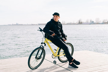 a young man in black sportswear stands on a pontoon near the lake and holds a yellow Bicycle near him on a Sunny spring day