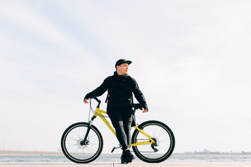 Fototapeta premium a young man in black sportswear stands on a pontoon near the lake and holds a yellow Bicycle near him on a Sunny spring day