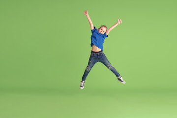 Jumping high. Happy boy playing and having fun on green studio background. Caucasian kid in bright...