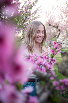 Portrait of young smiling woman in field or garden of beautiful spring trees in bloom. Feminine and sensual, natural beauty of blonde girl in pink flowers. Soft and tranquil pretty smile