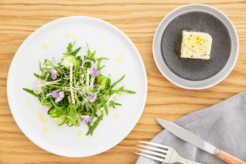 Arugula salad with bean sprout, enoki mushrooms, cucumber and edible flowers, feta cheese on the side over a wood background. Napkin, knife and fork. Flat lay. Vegetarian recipe.