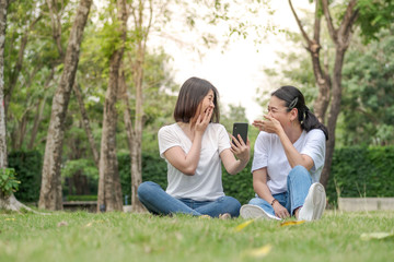 Fototapeta premium Two happy young girl friends sitting on grass in a park. Looking at smartphone.