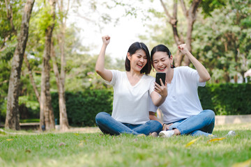 Two happy young girl friends sitting on grass in a park. Looking at smartphone.