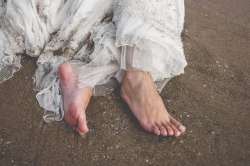 Bare feet of a bride with her messy dress on the beach.