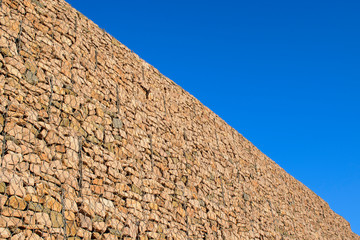 Fototapeta na wymiar High wall of gabions. Stones in a metal mesh. Blue sky background.