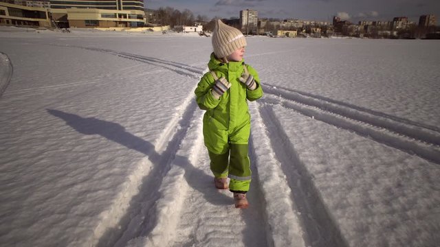 Elementary student in green snowsuit and carrying backpack walking along snow-covered road through field to school on winter day.