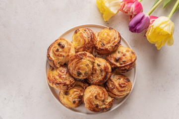 Homemade puff buns with custard cream and raisins in a ceramic plate on a light background. Nearby is a bouquet of colorful tulips. Copy space