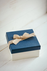 Square gift box with blue lid and burlap bow on white wooden background.