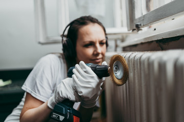 Beautiful and diligent middle age handy woman renovating her old home or apartment. She is holding professional grinder and brushing radiator for later painting. Do it yourself housework concept.
