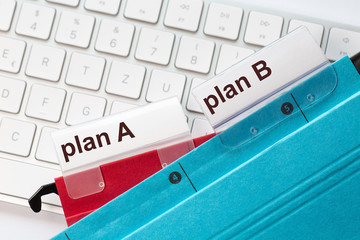 The  words plan A and plan B can be seen on the labels of a red and blue hanging folders. The hanging folders are on a computer keyboard.