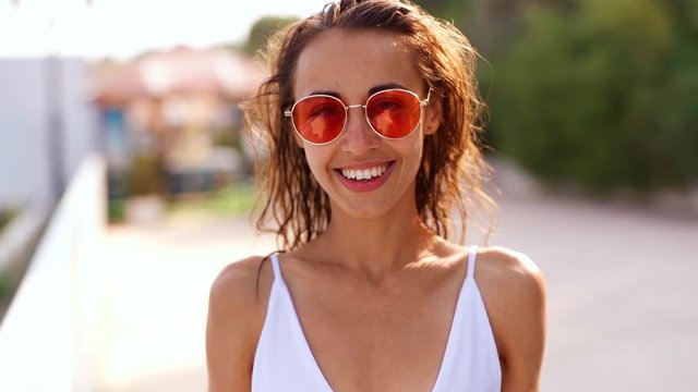 joyfully smiling Beautiful young tanned multyethnic woman in white bikini and pink sunglasses standing on bay at seaside on resort and smiling. slow motion footage