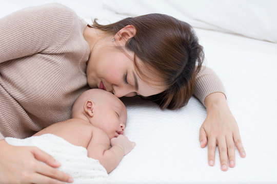 Beautiful mom is tenderly cuddles the newborn baby gently while the infant is sleeping on the white mattress. Asian mother lying near a baby and kissing on the head with love and care.