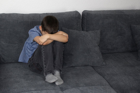 Child, Boy Sitting On A Gray Sofa In The Room, Clasping His Knees With His Hands And Covering His Face, The Concept Of Bad Mood And Education