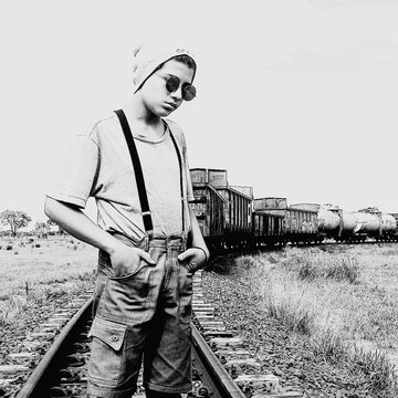 Boy In Bib Overalls Standing On Railroad Track Against Clear Sky