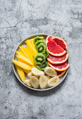 Fresh tropical fruits mango, kiwi, bananas, grapefruits in a plate on a grey background, top view. Delicious healthy dessert, snack