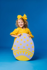 child a blonde girl in a yellow dress is smiling standing with a row of large decorative Easter eggs on a blue background