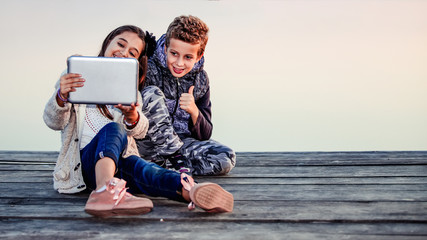 Two cute little friends, boy and girl having fun playing on digital tablet, celebrating success showing gesture thumb up