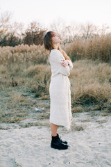 portrait of a young beautiful girl in a white dress, a warm knitted cardigan and black shoes among dry reeds and green bushes on the Bank of a river with white sand in the autumn time at sunset