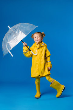 child girl blonde smiling in a yellow raincoat and rubber boots holding an umbrella stands on a blue background in the Studio, space for text