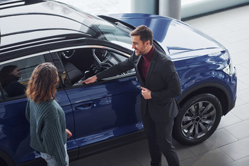 Fototapeta na wymiar Professional salesman assisting young girl by choosing new modern automobile indoors