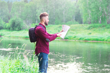 A man in a shirt and jeans with a backpack by the river looks forward and holds his hand on a map of the area