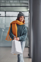 Smiling and feeling happy. Beautiful cheerful girl in yellow scarf and in warm clothes standing indoors with shopping bags in hands