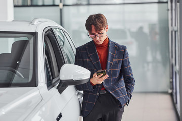 Hipster in formal wear and in eyewear standing near white modern car indoors