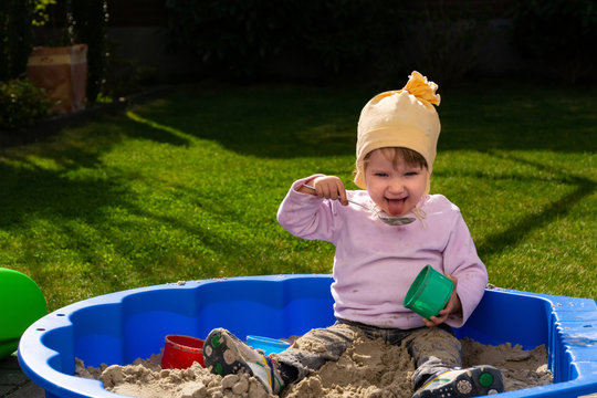A happy smiling kid plays in sandbox on a sunny day. A child using a spoon tries to taste sand with a tongue. Self-immunization concept.
