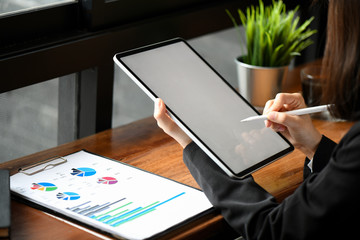 Cropped shot of a female employee using a pen to write on a tablet and graph documents on the table.