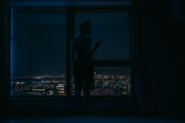 Silhouette of a young woman on the panoramic window background with the glass of vine in her hand and is looking on the night city landscape
