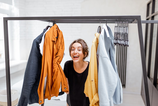 Portrait Of A Young And Cheerful Woman Choosing Casual Clothes To Wear, Standing Between Hangers And Looking Out Of The Clothes In The Wardrobe At The Bedroom