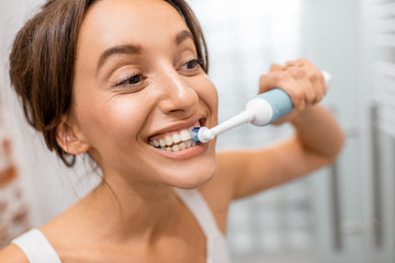 Young and cheerful woman brushing teeth with electric toothbrush during morning hygiene procedures in the bathroom, facial portrait