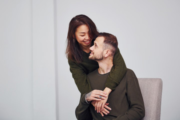 Happy multi ethnic couple in casual clothes embracing each other indoors in the studio. Caucasian guy with asian girlfriend