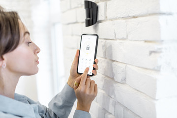 Woman controlling alarm system with smart phone and special mobile application wireless, standing...