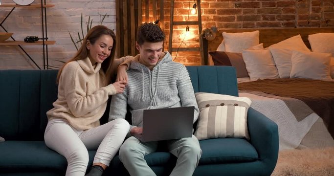 Young Couple spending Time in modern Loft Flat-Studio, surfing in the Internet. Spending Leisure together at Home, wearing Home Clothes. Cozy Evening. Nice Couple. Laptop Users. Love.