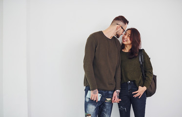 Cheerful multi ethnic couple with backpack and phone standing together indoors in the studio against white background