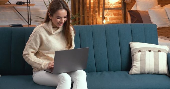 Young Girl looking happy Shocked while using her Laptop, becoming good News. Attractive Young Woman browsing Internet on Sofa in modern loft Flat. Wearing Home Outfit. Freelance. Working for Abroad.