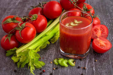 Tomato juice in a glass Cup , celery stalks , a mix of peppers, sliced and whole tomatoes on a dark wooden table.
