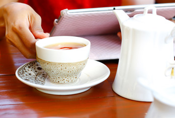 tea cup and teapot on wood table, one hand holding tea cup and another hand hold tablet 