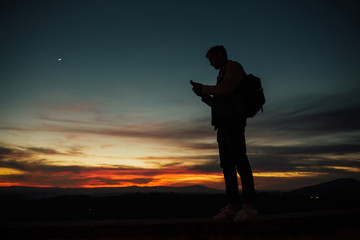 Silhouette of a man with backpack standing against the setting sun, checking messages on his phone, with a colorful clouds overhead. Young man holding mobile phone. 