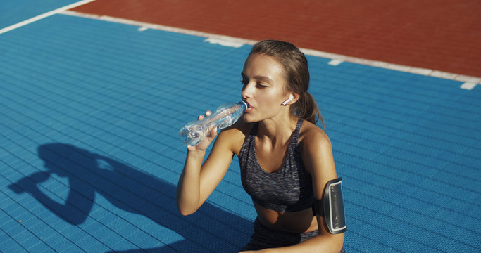 Pretty Caucasian young sporty woman sitting at sport court and drinking cold water from bottle after working out. Charming sportswoman resting and on sunny day after doing exercises outdoors.