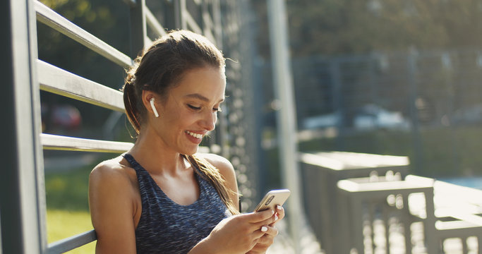 Pretty smiled Caucasian woman in sporty style and airpods tapping and texting message on smartphone on sunny warm day. Beautiful cheerful young sportswoman using phone for chatting at sport court.