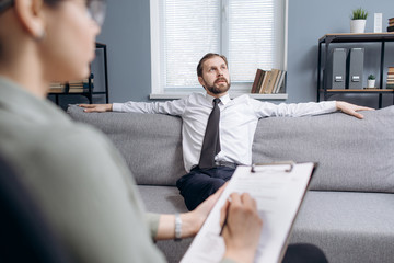 Relaxed bearded man in white shirt and black tie sitting comfortably on grey couch during therapist session. Mature patient visiting female doctor to get professional advice.