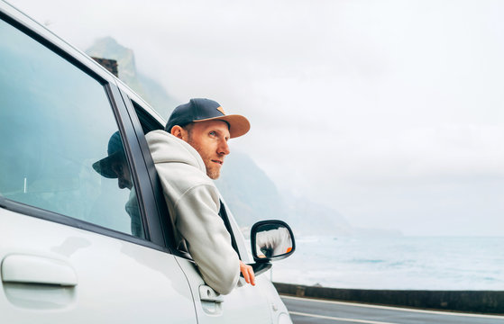 Man dressed warm hoodie and baseball cap driving his new car on the lonely road leading by the sea coast. Happy auto owner concept image.