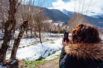 Beautiful curly brunette woman taking selfie portrait in the Pyrenees in winter, Spain. Mountains covered by snow, sunny day. girl video chatting on mobile phone sharing with friends family social