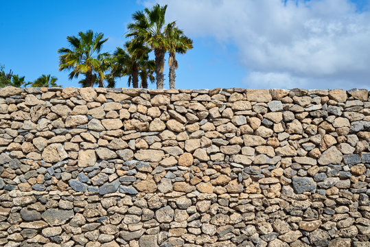 Beschaffenheit einer grauen und schwarzen Lava-Natursteinmauer als abstrakter Hintergrund mit Himmel und Palmen im Hintergrund