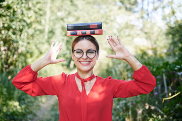 Fototapeta na wymiar Young blond woman, wearing red shirt and eyeglasses, holding two books on her head. Student with black and red books, smiling. Studying fun. Pretty girl, balancing with books on her head,