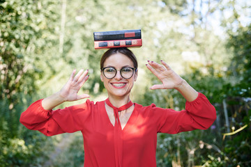 Fototapeta na wymiar Young blond woman, wearing red shirt and eyeglasses, holding two books on her head. Student with black and red books, smiling. Studying fun. Pretty girl, balancing with books on her head,