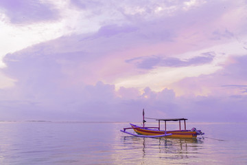 Traditional Balinese wooden fishing boat at dawn on Sanur beach in Bali. Cloudy pink dawn sky. Reflection in the water. Calm at sea